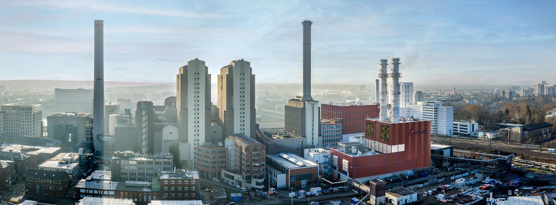 Luftaufnahme eines Industrieareals mit Kraftwerksgebäuden und Schornsteinen, dahinter Stadt und Bahntrassen.