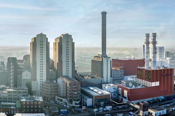 Luftaufnahme eines Industrieareals mit Kraftwerksgebäuden und Schornsteinen, dahinter Stadt und Bahntrassen.