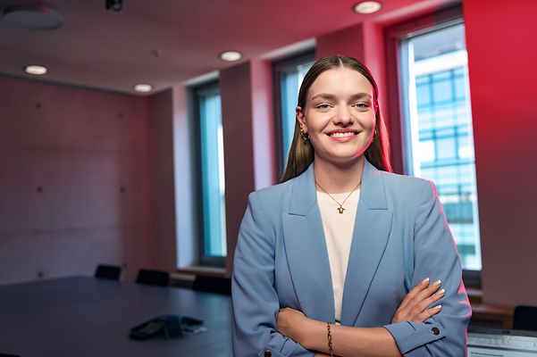 Frau mit verschränkten Armen lächelt in einem Meeting Raum in die Kamera 