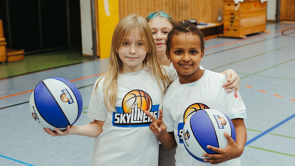 Drei Mädchen stehen in einer Halle mit Basketbällen und lächeln in die Kamera