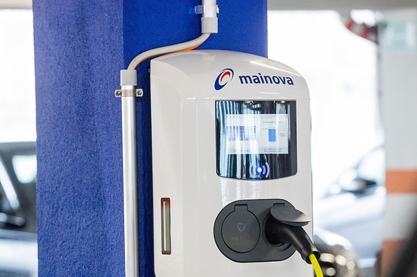 Mainova Ladestation im Parkhaus, blau-weiße Umgebung, Kabel an der Wand, im Hintergrund ein Auto.