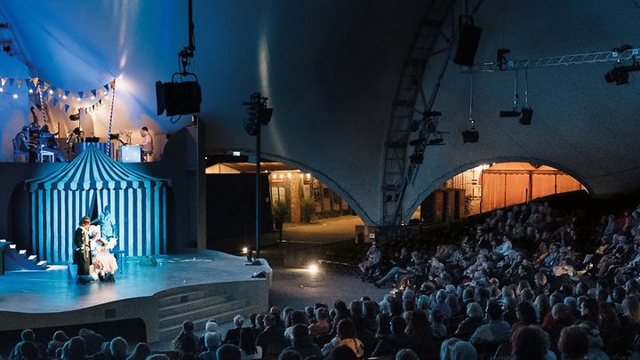 Freilichtbühne mit Theaterstück, beleuchtete Kulisse und viele Zuschauende unter großem Zeltdach