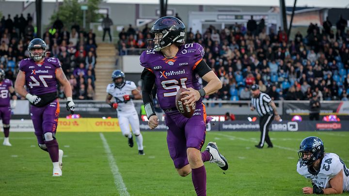 Footballspieler in lila Galaxy Trikot läuft mit Ball über Spielfeld, Gegenspieler am Boden und Publikum im Stadion dahinter.