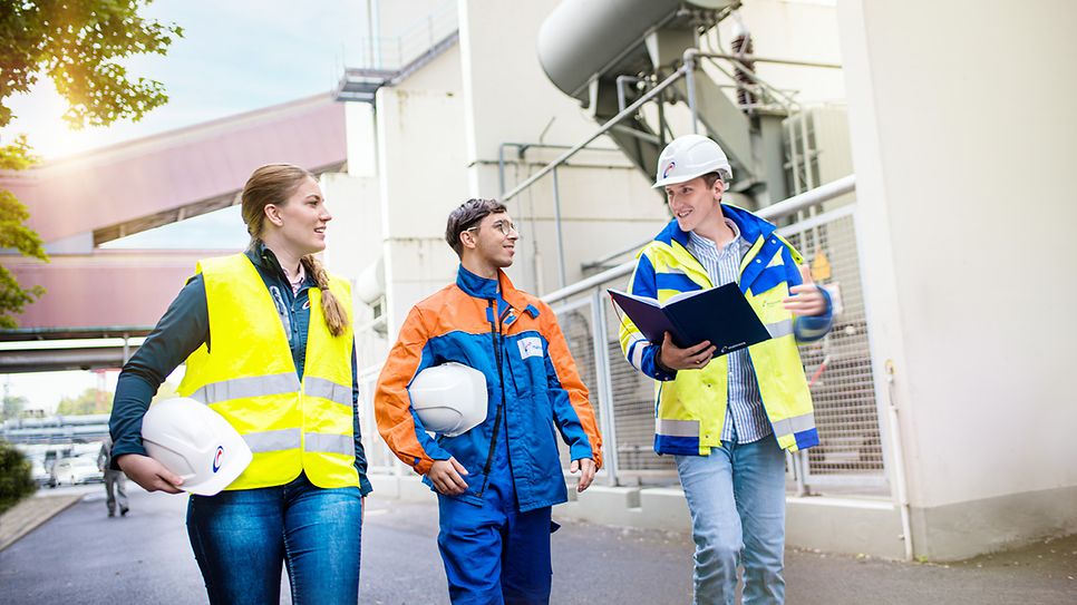 Drei Mitarbeitende mit Schutzhelmen und Warnkleidung besprechen sich auf dem Gelände einer Industrieanlage.