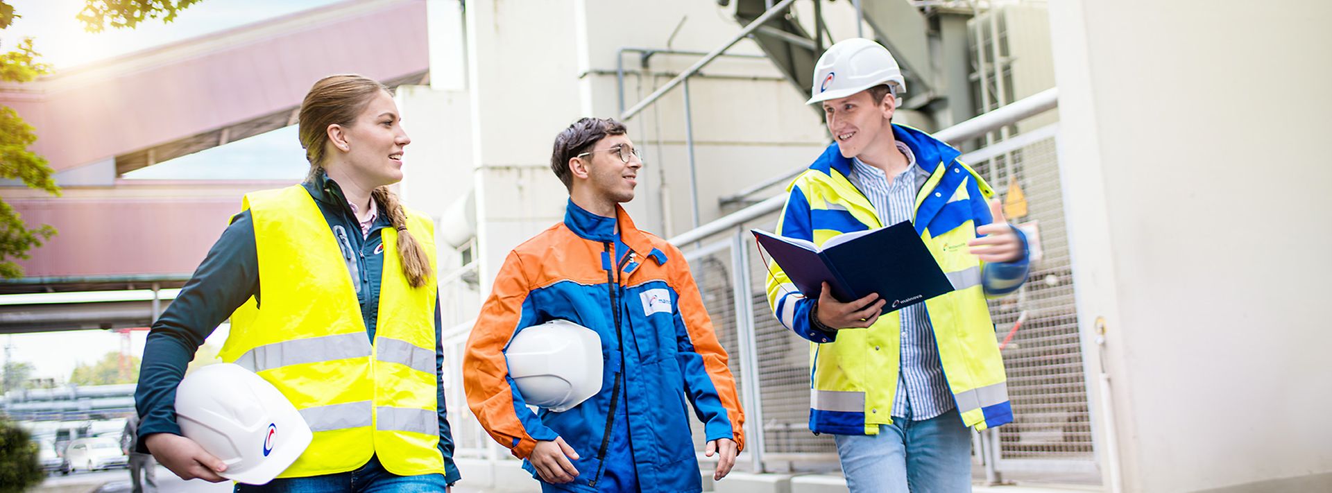 Drei Mitarbeitende mit Schutzhelmen und Warnkleidung besprechen sich auf dem Gelände einer Industrieanlage.