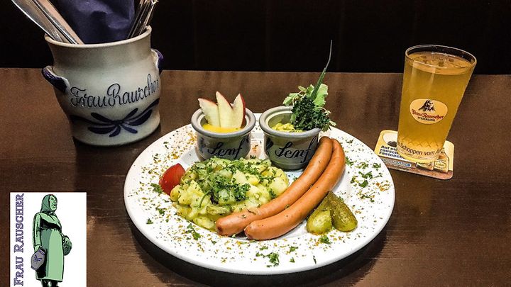 Frankfurter Würstchen mit Kartoffelsalat, Senf, Gurken und Apfelwein auf einem Tisch serviert.
