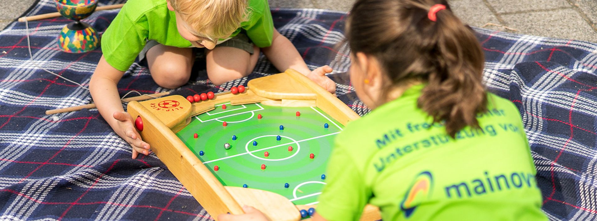 Junge und Mädchen in grünen Mainova T-Shirts spielen mit Tischfußball-Kasten auf einer Picknickdecke