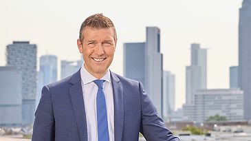 Mann in blauem Anzug mit blauer Krawatte lehnt an einem Geländer. Lächelnd vor einer modernen Stadtkulisse.