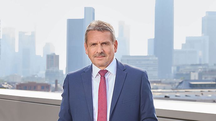 Mann im blauen Anzug mit roter Krawatte steht auf einer Terrasse vor einer Skyline. Professioneller, formeller Ausdruck.