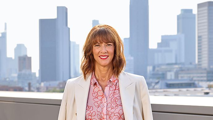 Frau in weißem Blazer mit rosa Bluse und Muster, stehend vor Skyline. Freundlicher Gesichtsausdruck.