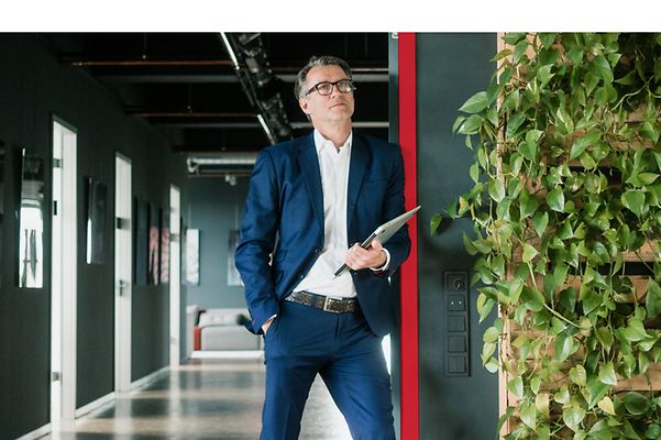 Geschäftsmann lehnt in Gebäudeeingang, links und rechts von ihm ranken sich Grünpflanzen an den Wänden