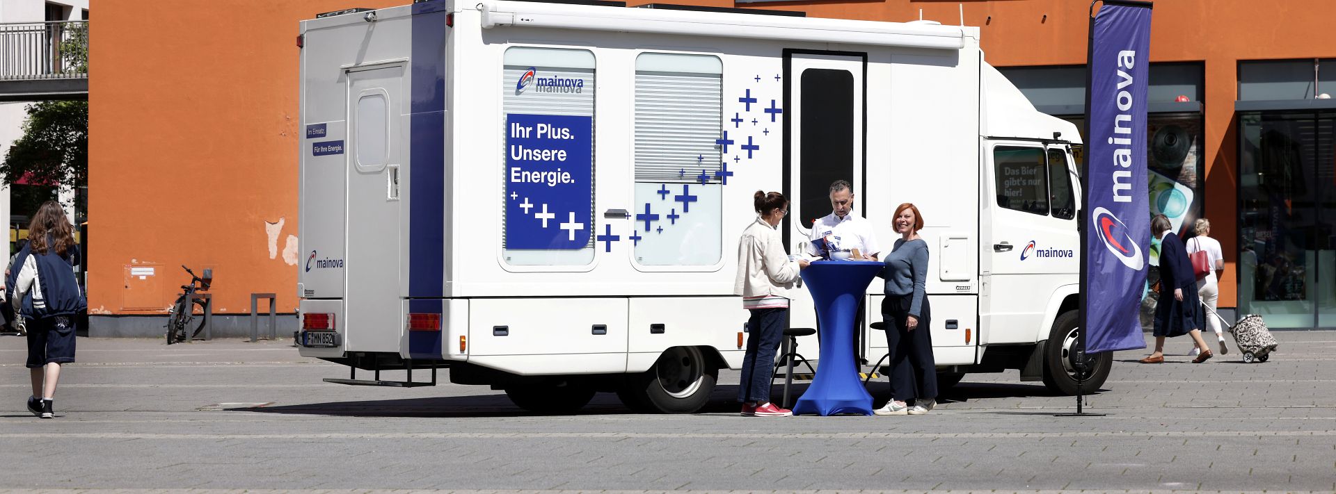 © Mainova Infomobil der Mainova mit blauem Stehtisch, drei Personen im Gespräch und orangem Gebäude im Hintergrund.