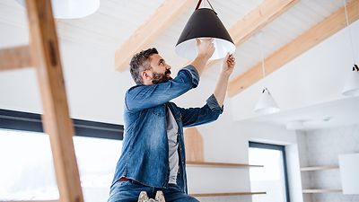 © stock.adobe.com/Halfpoint Mann sitzt auf einer Leiter und schraubt eine LED-Lampe in eine Deckenleuchte