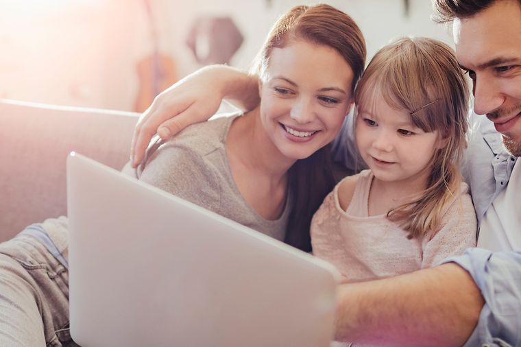 © iStock.com/Geber86 Frau und Mann mit kleiner Tochter sitzen eng zusammen auf dem Sofa und schauen auf einen Laptop