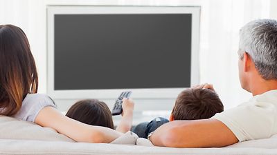 Rückansicht: Eltern und zwei Kinder auf der Couch, vor einem großem TV-Bildschirm