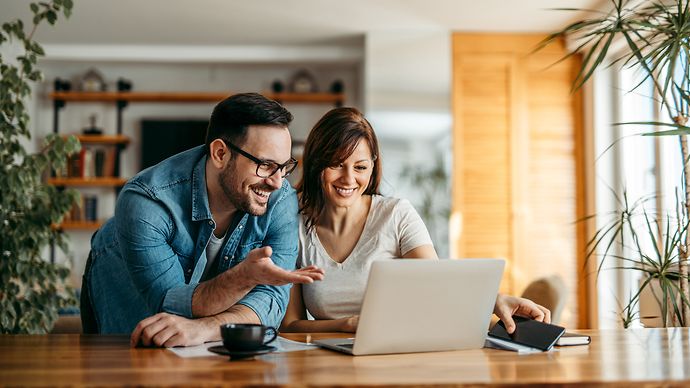 © Adobestock/bnenin Paar sitzt an einem Holztisch und schaut gemeinsam auf einen Laptop, umgeben von Pflanzen und gemütlicher Einrichtung.