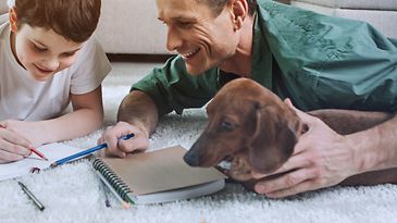 Vater, Sohn und Dackel liegen auf dem Wohnzimmerteppich und machen schriftliche Hausaufgaben