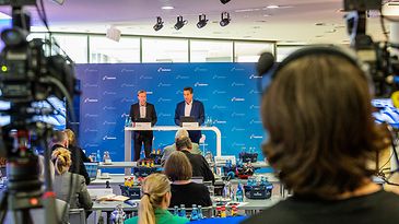 © mainova/DianaFunk Konferenz mit zwei Sprechern auf der Bühne, umgeben von Kameras und Publikum in einem modernen Raum.