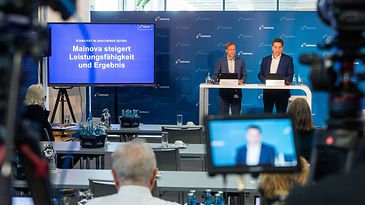 Zwei Personen bei Mainova-Pressekonferenz; Bildschirm mit Titel, Publikum sitzt, Kameras filmen.