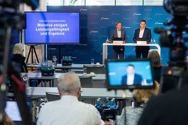 Zwei Personen bei Mainova-Pressekonferenz; Bildschirm mit Titel, Publikum sitzt, Kameras filmen.