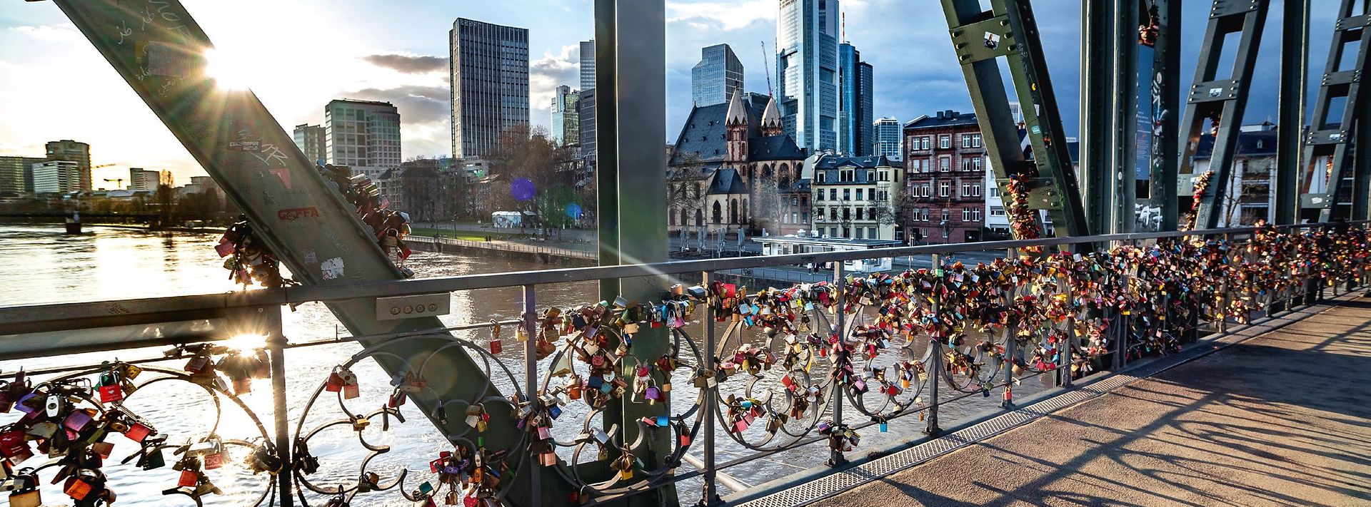 Liebesschlösser an einer Brücke über den Main, dahinter Frankfurter Skyline und Sonne im Abendlicht.