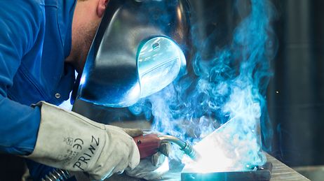 Ein Schweißer mit Schutzhelm und Handschuhen arbeitet an einem Metallstück, umgeben von blauem Rauch.