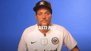 Ein Mann mit einem weißen Fußballtrikot und schwarzer Kappe vor blauem Hintergrund.