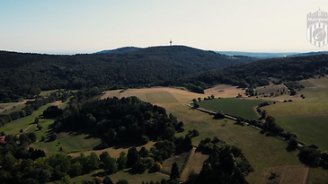 Luftaufnahme einer hügeligen Landschaft mit Feldern, Wäldern und einem Funkturm auf dem höchsten Hügel im Hintergrund.