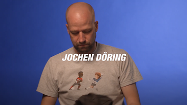 Eine Person mit Glatze vor blauem Hintergrund, trägt ein graues T-Shirt mit Comicfiguren.
