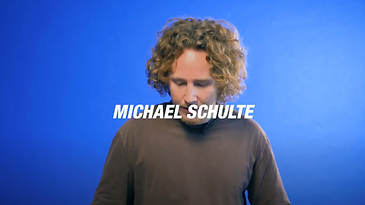 Eine Person mit lockigem Haar steht vor einem blauen Hintergrund.