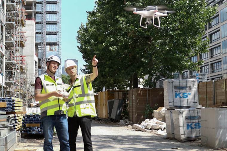 Zwei Bauarbeiter mit Warnwesten und Helmen steuern eine Drohne auf einer Baustelle.