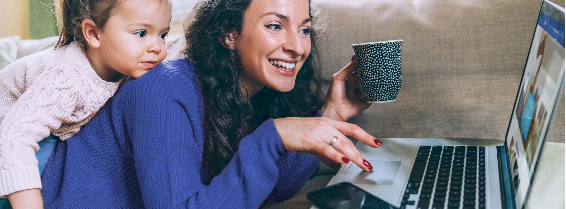 © iStock.com/filadendron Frau liegt auf der Couch und surft am Laptop mit Tochter auf dem Rücken