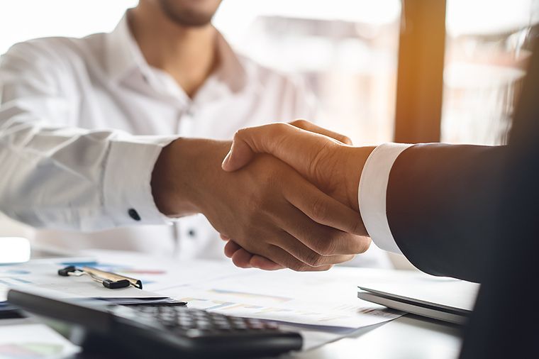 Zwei Personen in Businesskleidung geben sich über einem Schreibtisch mit Dokumenten und Taschenrechner die Hand.