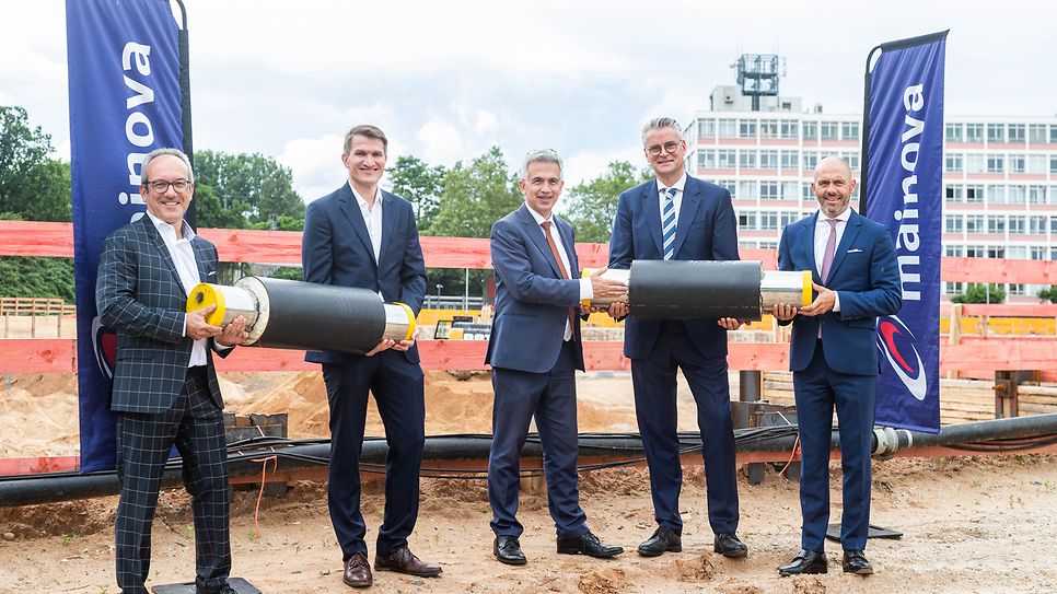 Fünf Männer in Anzügen halten Rohre bei einer Baustelle.