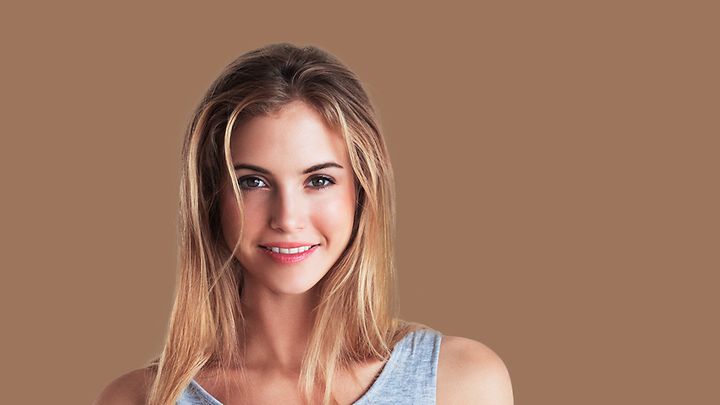 Eine Frau mit langen, blonden Haaren und einem grauen Oberteil vor einem braunen Hintergrund.