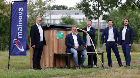 Fünf Männer in Anzügen stehen und sitzen vor einer Holzhütte im Freien, neben einem Mainova-Banner.