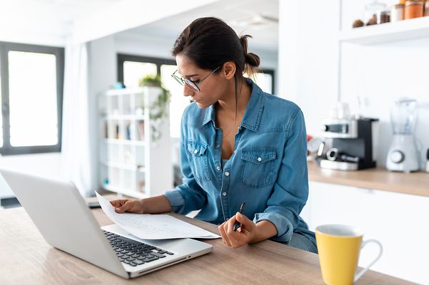 © stock.adobe.com/nenetus Junge Frau sitzt zu Hause vor ihrem Laptop und füllt einen Antrag aus.
