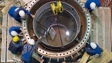 Sechs Arbeiter in Schutzkleidung installieren ein großes, zylindrisches Metallteil mit einem Kran in eine industrielle Anlage.