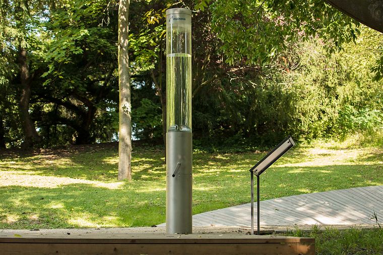 Ein silberner Zylinder mit einem durchsichtigen oberen Teil, der Wasser enthält, steht im Freien neben einem Schild.