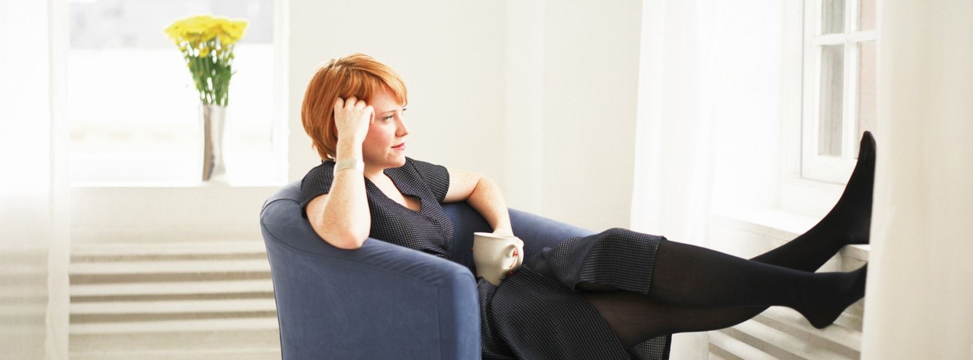 Junge Frau mit Kaffeetasse sitzt in einem Sessel und hat die Füße auf der Fensterbank.