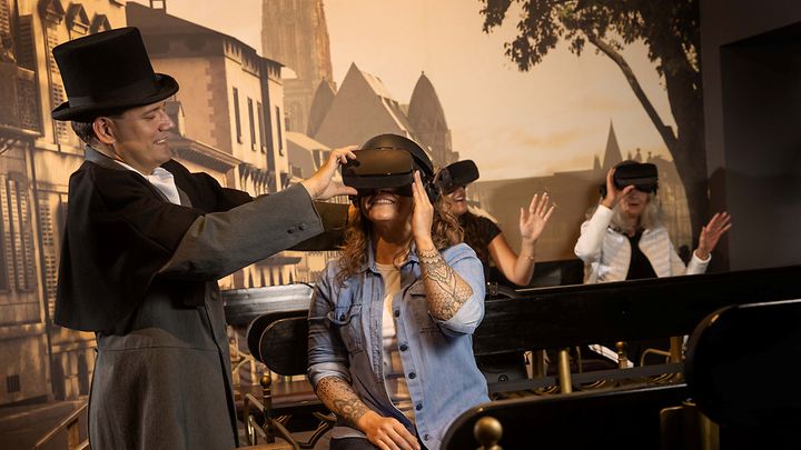 3 Personen sitzen sich gegenüber auf Bänken und haben VR Brillen auf