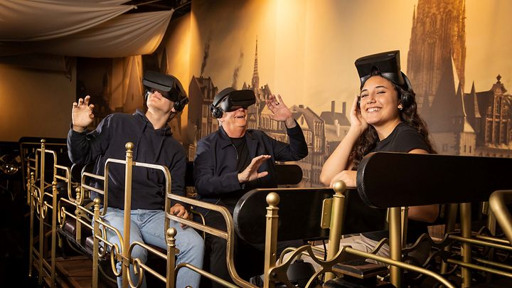 © TimeRide 3 Personen sitzen sich gegenüber auf Bänken und haben VR Brillen auf