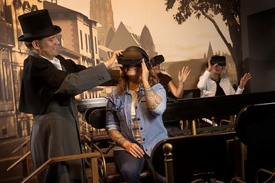 3 Personen sitzen sich gegenüber auf Bänken und haben VR Brillen auf