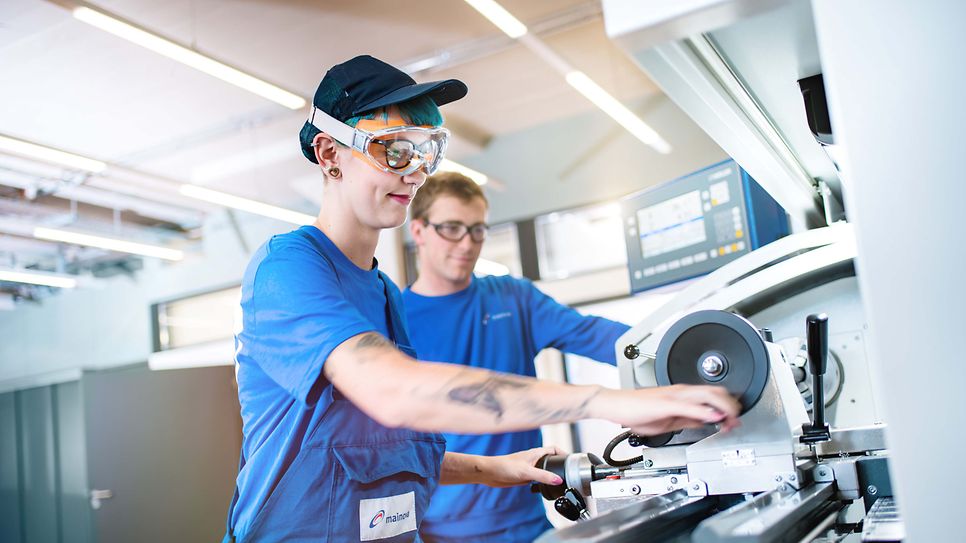 Junge Frau steht an Drehmaschine und der Ausbilder schaut der Arbeit zu.