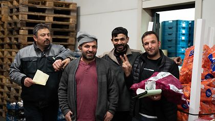Vier Männer stehen in einem Lagerraum, umgeben von Paletten und Säcken mit Orangen.