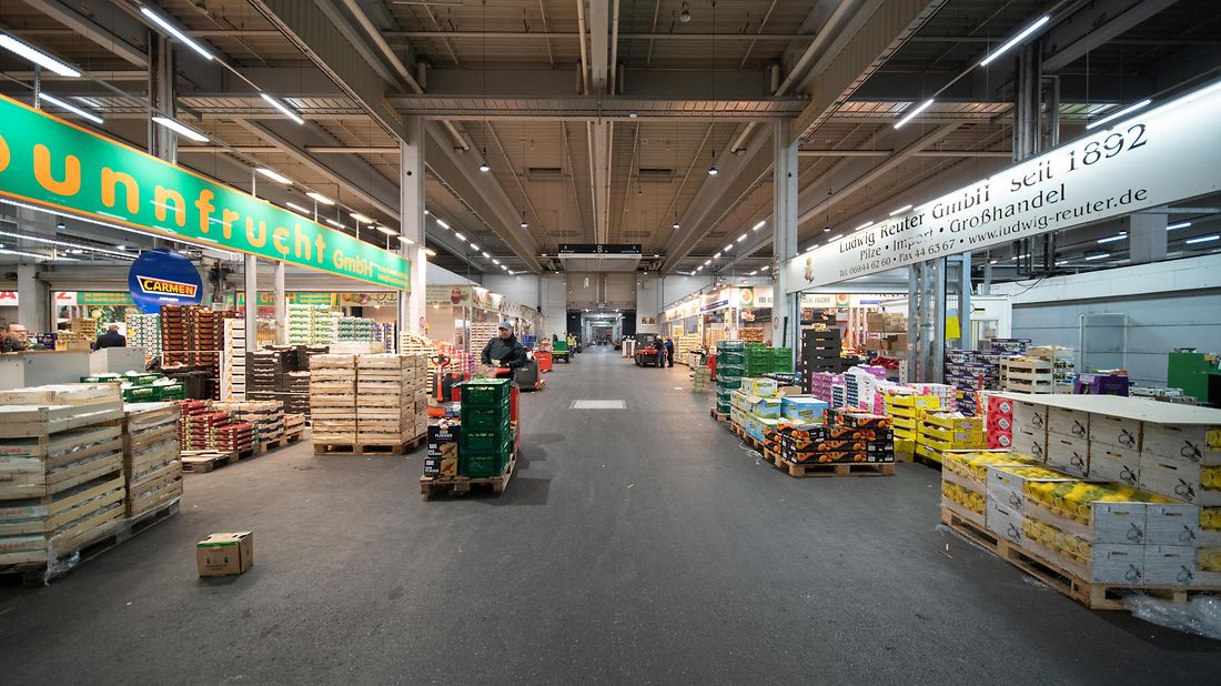 Ein großer, gut beleuchteter Marktgang mit gestapelten Kisten und Schildern für Obst und Gemüse.