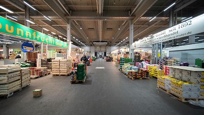 Ein großer, gut beleuchteter Marktgang mit gestapelten Kisten und Schildern für Obst und Gemüse.
