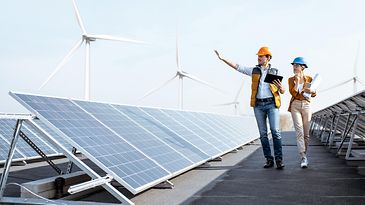 Geschäftsleute begutachten Photovoltaikanlage auf dem Dach eines Gebäudes mit Windrädern im Hintergrund
