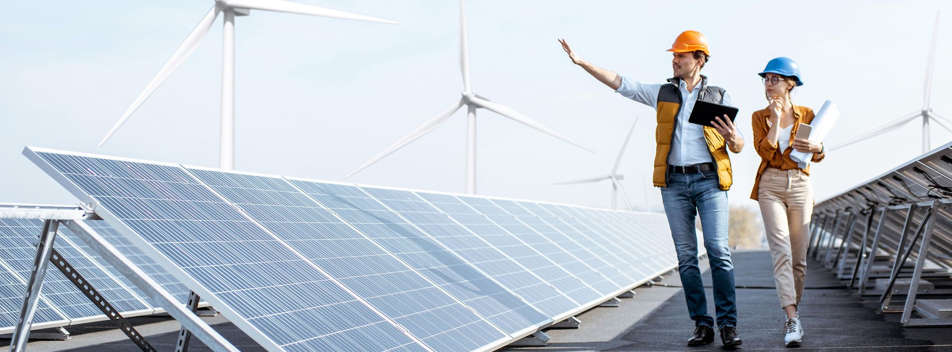 © stock.adobe.com/rh2010 Geschäftsleute begutachten Photovoltaikanlage auf dem Dach eines Gebäudes mit Windrädern im Hintergrund