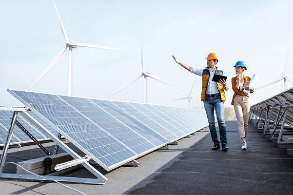 © stock.adobe.com/rh2010 Geschäftsleute begutachten Photovoltaikanlage auf dem Dach eines Gebäudes mit Windrädern im Hintergrund